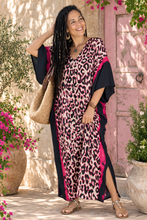 Load image into Gallery viewer, A woman wearing a pink and black leopard print kaftan dress with black wide sleeves, standing outdoors in front of a pink door and potted plants.