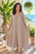 Load image into Gallery viewer, A woman stands on a sunny porch wearing a loose, knee-length kaftan dress with a beige and white geometric pattern. The background features a beautiful ocean view, potted plants, and a white railing.