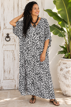 Load image into Gallery viewer, A woman wearing a black and white leopard print maxi dress with short wide sleeves, standing in front of a rustic white wooden door next to a large potted plant.