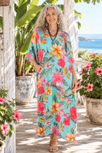 Load image into Gallery viewer, A smiling woman with curly gray hair stands on a sunlit porch wearing a flowing turquoise maxi dress with a bold tropical floral print in pink, orange, and green. Large potted plants and a glimpse of the ocean are visible in the background.