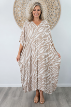 Load image into Gallery viewer, A smiling woman models a loose, V-neck kaftan dress with a beige and white zebra-like pattern, standing in front of a white wall with a decorative wooden wall art piece.
