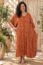 Load image into Gallery viewer, A woman wearing a rust-orange kaftan dress with a white abstract wave pattern, standing in front of a rustic wooden door and potted plants.
