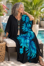Load image into Gallery viewer, A woman with curly gray hair sits on a patio chair by a swimming pool, wearing a black kaftan dress with a vibrant blue tropical palm leaf print. She is accessorized with a long beaded tassel necklace.
