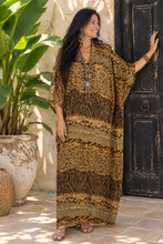 Load image into Gallery viewer, A woman wearing a bohemian-style  kaftan dress with zebra and leopard prints in brown, black, and gold, posing next to a rustic black door.
