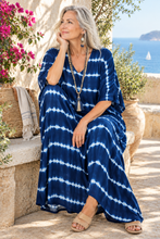 Load image into Gallery viewer, A woman with gray hair sits on a stone ledge outdoors by the sea, wearing a loose navy blue maxi dress with white tie-dye horizontal stripes. She is wearing beige sandals and a long beaded tassel necklace.
