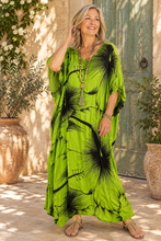 Load image into Gallery viewer, A woman wearing a bright lime green kaftan dress with a black floral print, standing in front of a rustic green door and potted plants.
