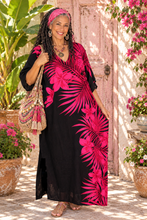 Load image into Gallery viewer, A woman wearing a black kaftan dress with a vibrant hot pink tropical floral and palm leaf print, styled with a matching pink headband, a woven tassel shoulder bag, and sandals, standing in front of a rustic pink wooden door with potted plants.

