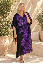 Load image into Gallery viewer, A woman wearing a black and purple tropical print kaftan dress, holding a woven straw tote bag, standing in a sunny outdoor patio setting with potted plants.
